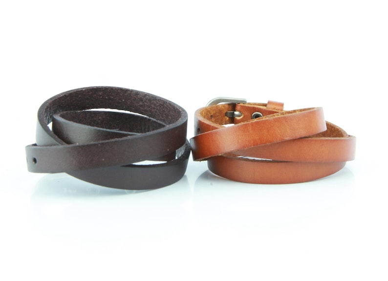 May include: Two leather wrap bracelets. One is dark brown, the other is tan. The dark brown bracelet is a simple wrap with a single hole. The tan bracelet has a silver buckle.