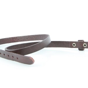 May include: A slender, dark brown leather belt with a silver buckle. The belt features multiple holes for adjustment and is designed for waist wear. The belt is made from a smooth, durable material, and is approximately 1.5 cm wide.