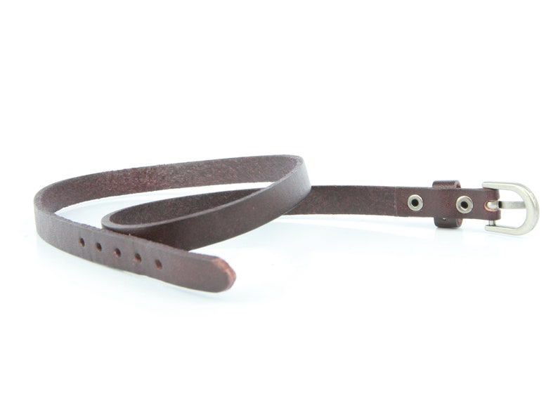 May include: A slender, dark brown leather belt with a silver buckle. The belt features multiple holes for adjustment and is designed for waist wear. The belt is made from a smooth, durable material, and is approximately 1.5 cm wide.