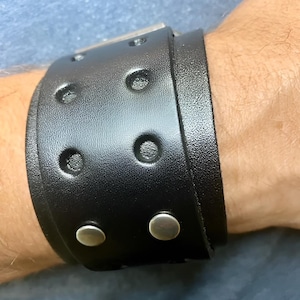 Puede incluir: Una pulsera ancha de cuero negro con remaches plateados y una hebilla rectangular. La pulsera tiene hendiduras circulares. Dos broches plateados aseguran la pulsera.