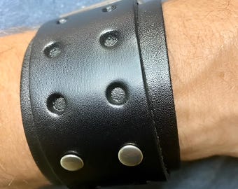 Könnte beinhalten: Ein breites, schwarzes Lederarmband mit silberfarbenen Nieten und einer rechteckigen Schnalle. Das Armband hat kreisförmige Vertiefungen. Zwei silberfarbene Druckknöpfe sichern das Band.