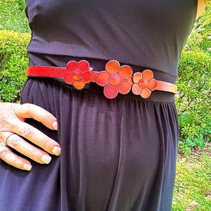 May include: A red leather belt with three large flower-shaped buckles. The belt is worn over a black dress.