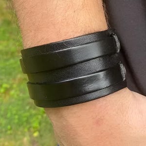 K&ouml;nnte beinhalten: Ein breites schwarzes Lederarmband mit drei &uuml;bereinanderliegenden B&auml;ndern. Das Armband wird am Handgelenk getragen, und das Leder scheint glatt und gl&auml;nzend zu sein. Die Manschette wird mit einer Schnalle befestigt.