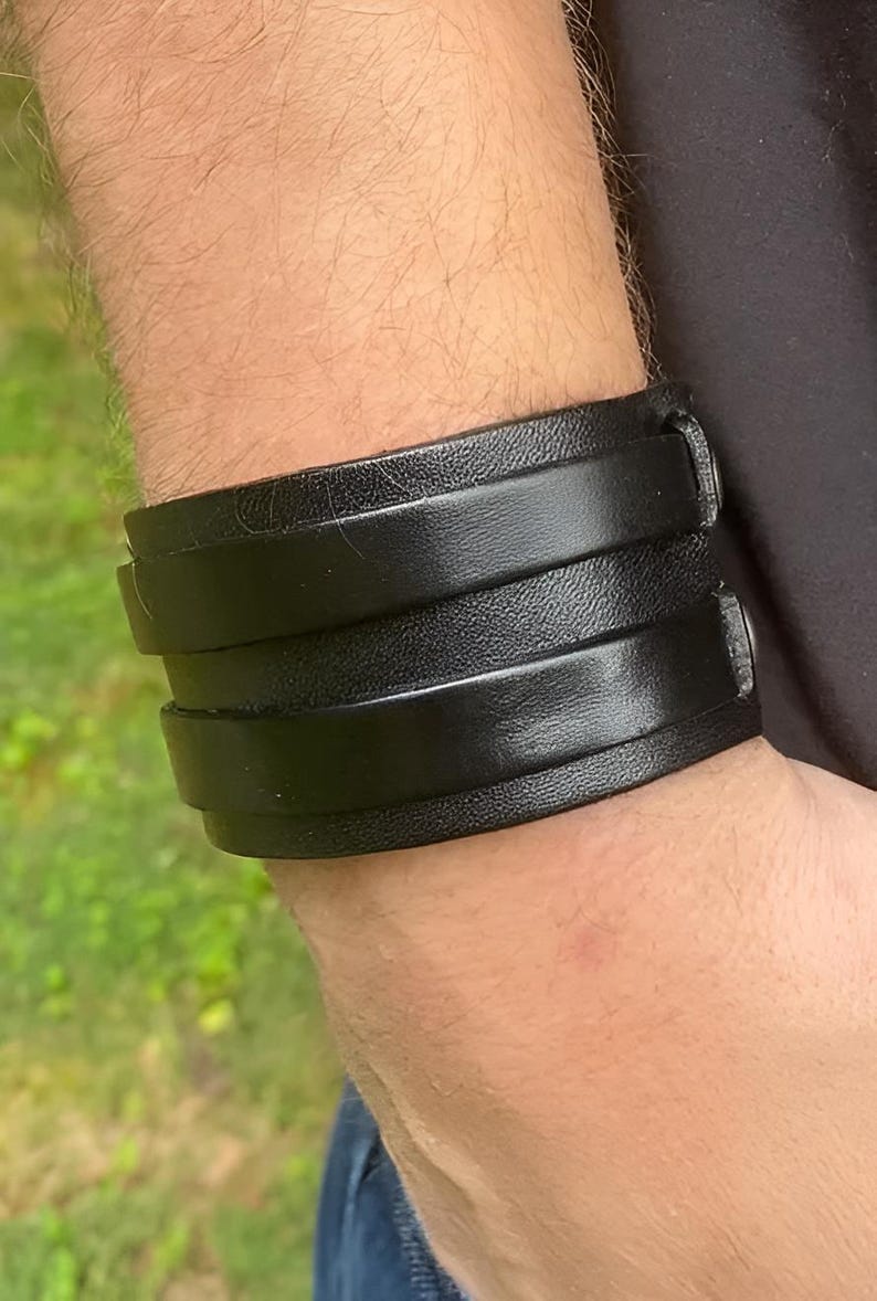 K&ouml;nnte beinhalten: Ein breites schwarzes Lederarmband mit drei &uuml;bereinanderliegenden B&auml;ndern. Das Armband wird am Handgelenk getragen, und das Leder scheint glatt und gl&auml;nzend zu sein. Die Manschette wird mit einer Schnalle befestigt.