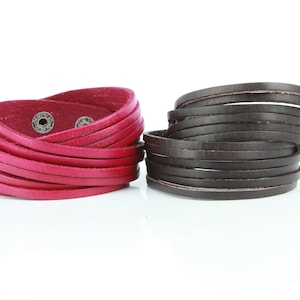 May include: Two leather wrap bracelets. One is a vibrant red, secured with silver snaps. The other is a deep brown. Both bracelets are made of multiple thin leather strips, creating a layered effect. The bracelets are displayed against a white background.