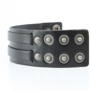 K&ouml;nnte beinhalten: Schwarzes Lederarmband mit zwei Riemen und silberfarbenen Metall-Druckkn&ouml;pfen und dekorativen Nieten. Das Armband ist gebogen und scheint ungetragen zu sein. Das Armband ist ein breiter Manschettenstil.