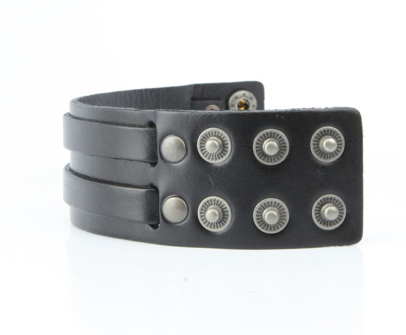 K&ouml;nnte beinhalten: Schwarzes Lederarmband mit zwei Riemen und silberfarbenen Metall-Druckkn&ouml;pfen und dekorativen Nieten. Das Armband ist gebogen und scheint ungetragen zu sein. Das Armband ist ein breiter Manschettenstil.