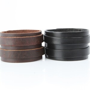 K&ouml;nnte beinhalten: Zwei Lederarmb&auml;nder. Eines ist braun, das andere ist schwarz. Jedes Armband besteht aus mehreren gestapelten Lederb&auml;ndern. Das braune Armband hat einen Used-Look, w&auml;hrend das schwarze Armband eine glatte Oberfl&auml;che hat.