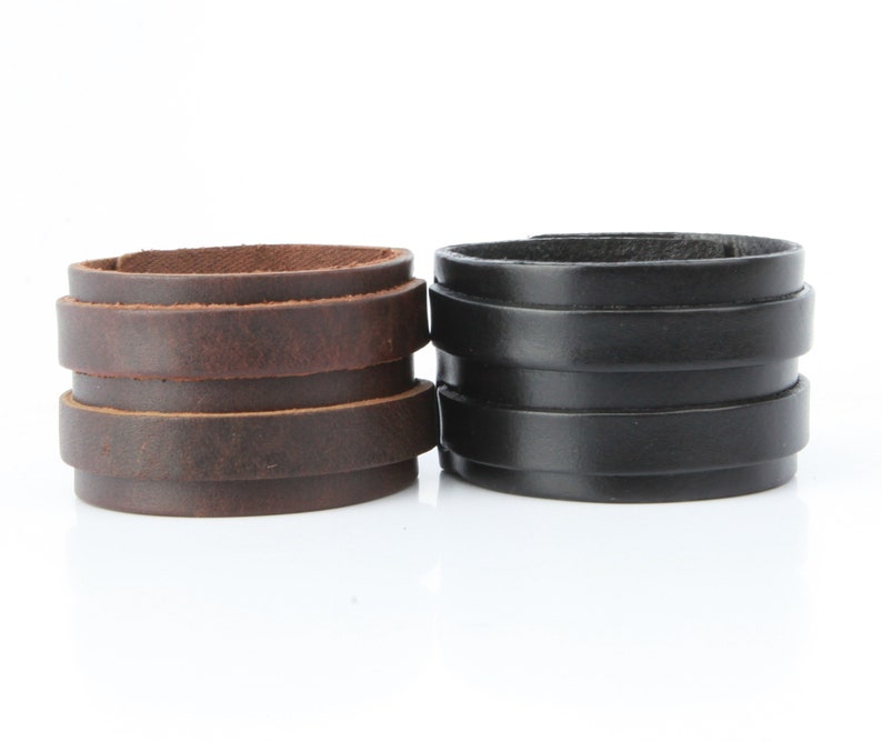 K&ouml;nnte beinhalten: Zwei Lederarmb&auml;nder. Eines ist braun, das andere ist schwarz. Jedes Armband besteht aus mehreren gestapelten Lederb&auml;ndern. Das braune Armband hat einen Used-Look, w&auml;hrend das schwarze Armband eine glatte Oberfl&auml;che hat.