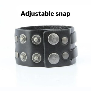 K&ouml;nnte beinhalten: Schwarzes Lederarmband mit silberfarbenen Nieten und Druckkn&ouml;pfen. Das Armband hat den Text "THS" auf den Druckkn&ouml;pfen und die Worte "Adjustable snap" stehen dar&uuml;ber.