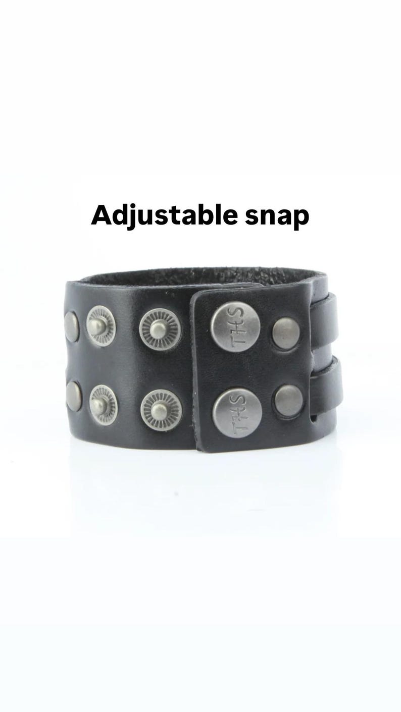 K&ouml;nnte beinhalten: Schwarzes Lederarmband mit silberfarbenen Nieten und Druckkn&ouml;pfen. Das Armband hat den Text "THS" auf den Druckkn&ouml;pfen und die Worte "Adjustable snap" stehen dar&uuml;ber.