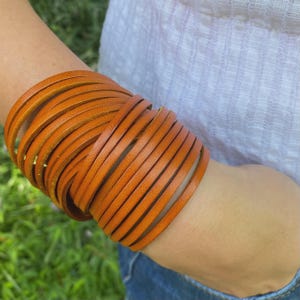 May include: A stack of orange leather strips wrapped around a wrist, forming a wide bracelet. The leather has a smooth texture and a warm, earthy tone. The bracelet is worn against a background of a white shirt and blue jeans.