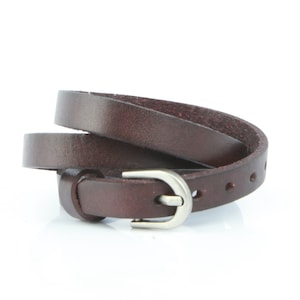 May include: A dark brown leather belt with a silver buckle. The thin belt has several holes for adjustment. The buckle is a simple, rounded design. The belt is coiled, showing its length.