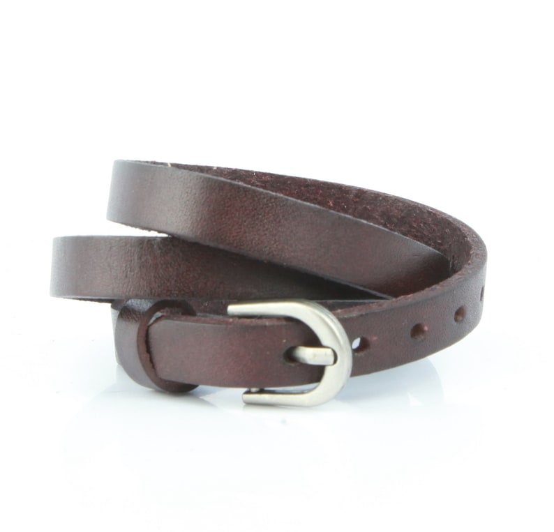 May include: A dark brown leather belt with a silver buckle. The thin belt has several holes for adjustment. The buckle is a simple, rounded design. The belt is coiled, showing its length.