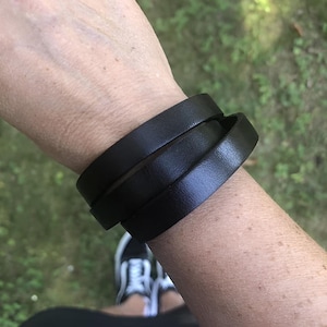 Könnte beinhalten: Schwarzes Lederarmband, das um ein Handgelenk gewickelt ist. Das Armband besteht aus drei Lederbändern. Der Hintergrund ist verschwommen, mit grünem Gras und einem Paar weißer Turnschuhe.