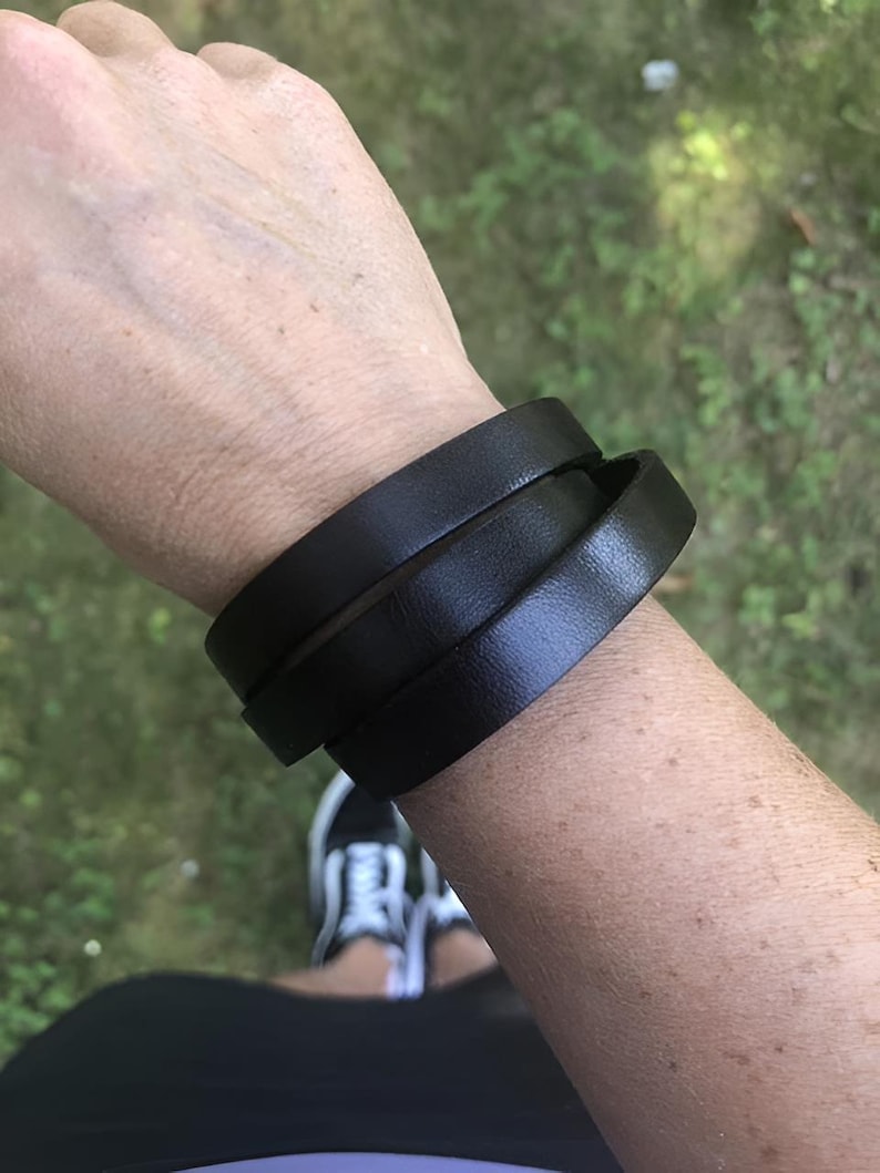 May include: A black leather bracelet wrapped around a wrist. The bracelet is made of three bands of leather. The background is blurred, with green grass and a pair of white trainers visible.