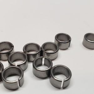 Pode incluir: Um grupo de 11 anéis de metal prateado com uma fenda em um lado. Os anéis estão dispostos em um grupo sobre uma superfície branca.