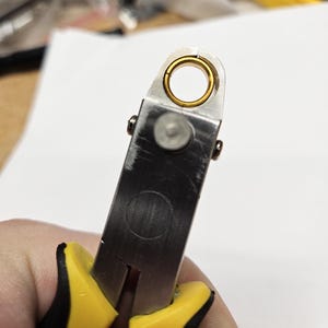 Puede incluir: Primer plano de unos alicates de joyería que sostienen un pequeño anillo de color dorado. Los alicates son de metal plateado con mangos amarillos y negros. El anillo es circular y tiene una superficie lisa.