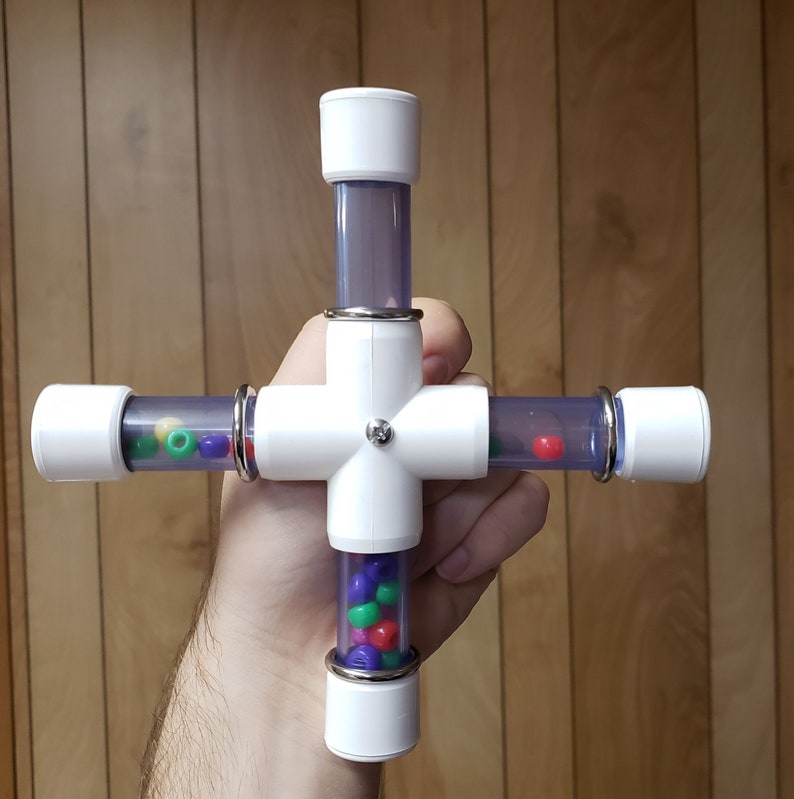 Puede incluir: Un juguete de pl&aacute;stico blanco en forma de cruz con cuatro tubos transparentes llenos de cuentas de colores. Las cuentas son rojas, verdes, moradas y blancas.