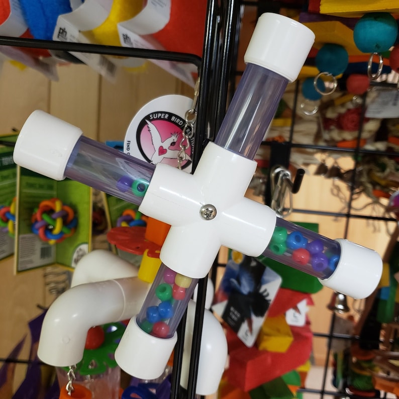 Puede incluir: Juguete para p&aacute;jaros de pl&aacute;stico blanco con cuatro tubos transparentes llenos de cuentas de colores. El juguete est&aacute; unido a una cadena de metal y tiene una etiqueta que dice "Super Bird Find Your Fun".