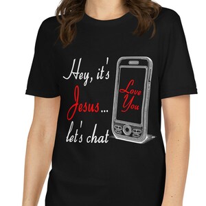 以下が含まれることがあります： 「Hey, it's Jesus... let's chat」の文字が白と赤の筆記体で書かれた黒いTシャツ。画面に「Love You」と書かれた銀色のフリップフォンのグラフィックも特徴です。Tシャツは人が着ています。