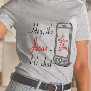 以下が含まれることがあります： 「Hey, it's Jesus... let's chat」のテキストと携帯電話のイラストが描かれたライトグレーのTシャツ。携帯電話の画面には「Love You」と表示されています。このシャツはクルーネックで半袖です。