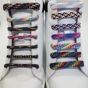 May include: White sneakers with colorful braided shoelaces. The shoelaces feature various patterns, including rainbow, pink, purple, and black and white designs. The laces are threaded through metal eyelets.