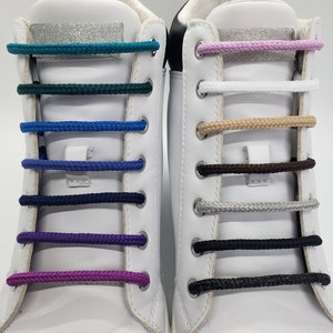 Op de afbeelding: Twee witte sneakers met verschillende gekleurde veters. De veters zijn in verschillende kleuren, waaronder turquoise, groen, blauw, paars, wit, beige, bruin, grijs en zwart.