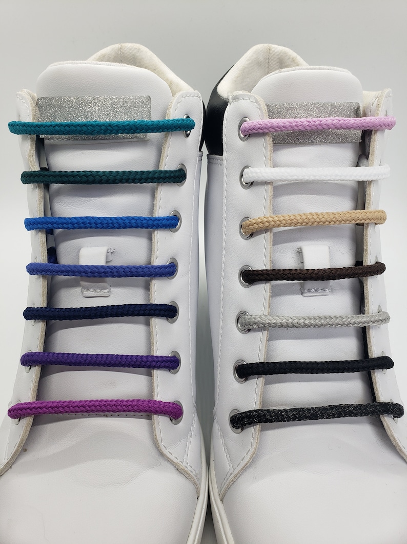 Op de afbeelding: Twee witte sneakers met verschillende gekleurde veters. De veters zijn in verschillende kleuren, waaronder turquoise, groen, blauw, paars, wit, beige, bruin, grijs en zwart.