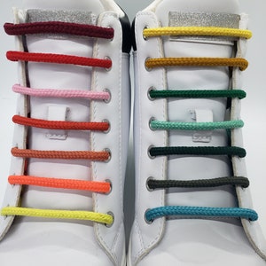 Op de afbeelding: Twee witte high-top sneakers met verschillende gekleurde veters. De veters zijn in een regenboogpatroon, waarbij elk paar veters een andere kleur heeft. De veters zijn bovenaan de schoen in een strik geknoopt.