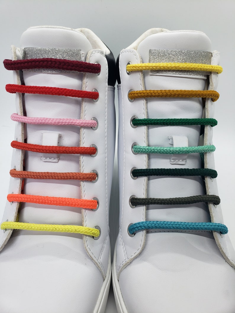 Op de afbeelding: Twee witte high-top sneakers met verschillende gekleurde veters. De veters zijn in een regenboogpatroon, waarbij elk paar veters een andere kleur heeft. De veters zijn bovenaan de schoen in een strik geknoopt.