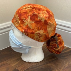 Puede incluir: Un gorro de quirófano con estampado de hojas otoñales en tonos naranja y dorado, que incluye una bolsa trasera fruncida y una cinta marrón. Se muestra sobre un maniquí blanco que lleva una mascarilla desechable de color azul claro.