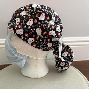 May include: A black scrub cap featuring a festive pattern of white ghosts wearing Santa hats, reindeer antlers, candy canes, and snowflakes. The cap has a gathered back section with white ties and is displayed on a mannequin head wearing a light blue mask.
