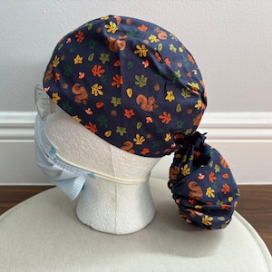 Pode incluir: Uma touca cirúrgica azul-marinho com um padrão repetitivo de esquilos castanhos, folhas de outono laranja e amarelas e pequenos cogumelos vermelhos. A touca possui uma secção traseira franzida com um fecho de atar para cobrir o cabelo.