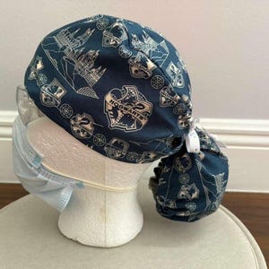 May include: A dark blue scrub cap featuring a repeating white pattern of the Hogwarts crest, castle silhouettes, and circular motifs. The cap has a gathered back with a white tie closure and is worn over a light blue disposable face mask.