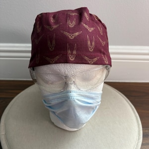May include: A maroon scrub cap featuring a repeating gold-colored pattern of winged spheres. The cap is worn on a white mannequin head that is also wearing clear protective safety glasses and a light blue disposable pleated face mask.