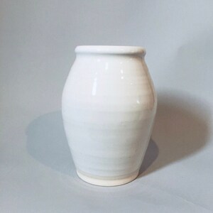 May include: A white ceramic vase with a rounded body and a slightly flared rim. The vase has a smooth, glossy finish and a subtle, uneven texture.