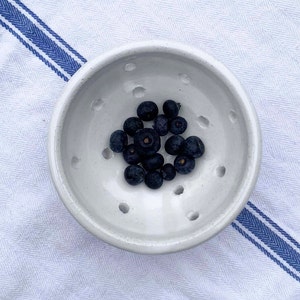May include: A white ceramic bowl with holes in the bottom, filled with blueberries. The bowl is sitting on a white and blue striped tablecloth.