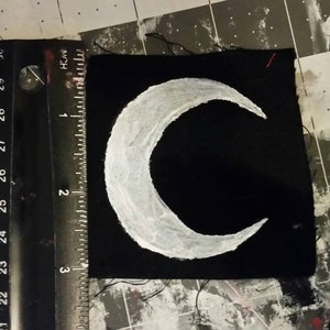 Könnte beinhalten: Ein schwarzer Stoff-Patch mit einem weißen Halbmond-Design. Der Patch ist etwa 5 cm breit und 5 cm hoch.