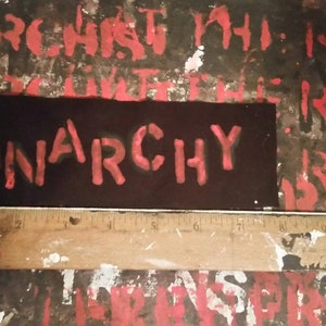 May include: A black patch with the word "ANARCHY" in red, block letters. The patch is approximately 10 cm long. The background is a distressed surface with red painted text.