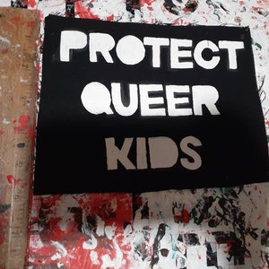 May include: A black square with the words "PROTECT QUEER KIDS" in bold white letters. The sign is on a surface with paint splatters.