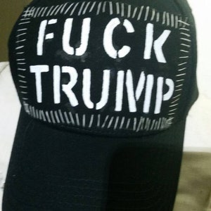 May include: Black baseball cap with the words "FUCK TRUMP" in white block letters on a rectangular patch. The patch is stitched onto the cap with white thread.