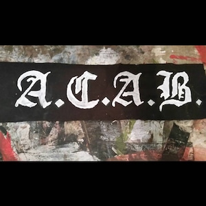 Peut inclure: Écusson rectangulaire noir avec les lettres blanches "A.C.A.B." en police gothique. Le patch est posé sur une surface aux couleurs variées, notamment rouge, blanc et gris.