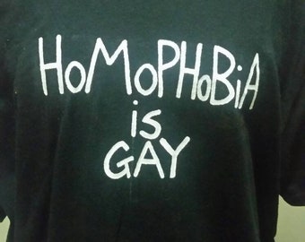 May include: Black t-shirt with the white text "HoMoPhObIa is GaY". The shirt has a crew neck and short sleeves. A white undershirt is visible at the neckline.