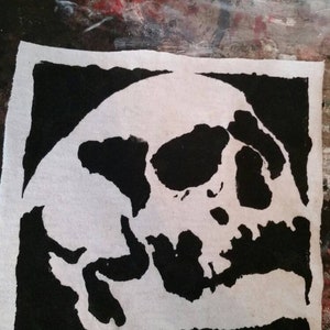 May include: A square, white fabric with a black skull design. The skull is depicted in a bold, graphic style, with the black areas defining the shape against the white background. The fabric has a slightly textured appearance.