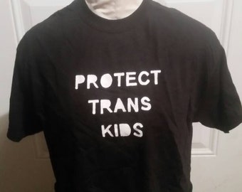 May include: Black t-shirt with the white text "PROTECT TRANS KIDS". The shirt is displayed on a mannequin. The mannequin is on a white background.
