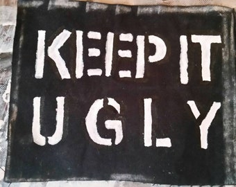 Pode incluir: Tela preta com as palavras "KEEP IT UGLY" estampadas em branco. O texto está centrado na tela, com uma aparência ligeiramente desgastada e feita à mão. O fundo é uma mistura de respingos de tinta.