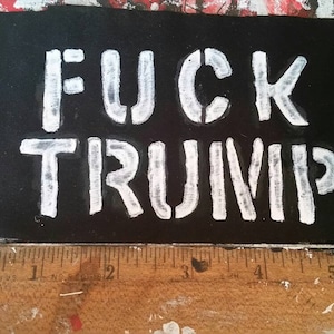 Può includere: Un rettangolo nero con le parole "FUCK TRUMP" dipinte in bianco. L'immagine include un righello di legno lungo il bordo inferiore e lo sfondo è un mix di vernice rossa, bianca e nera.