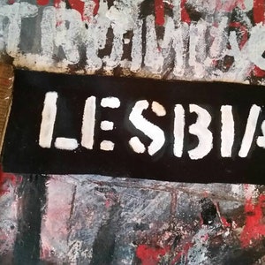 May include: A black rectangular patch with the word "LESBIAN" in white, stencil-style lettering. The patch is placed on a textured surface with red, white, and black paint. A wooden ruler is partially visible.