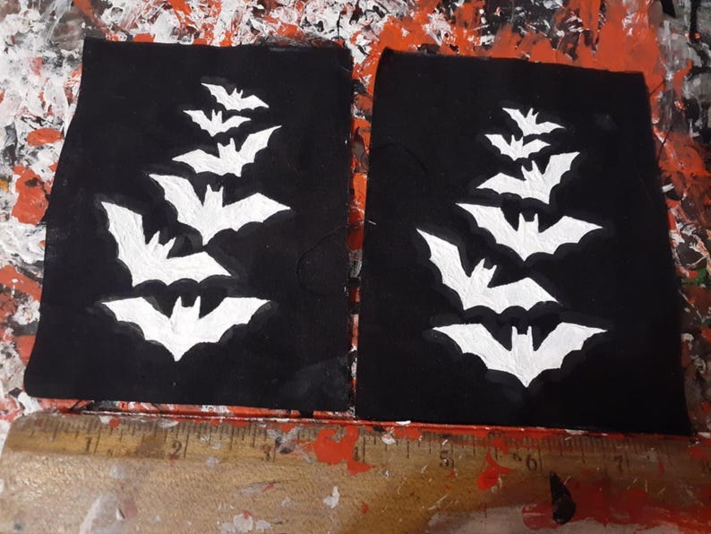 May include: Two black fabric patches with white painted bats in a cascading pattern. The bats are of varying sizes and are arranged in a vertical line.